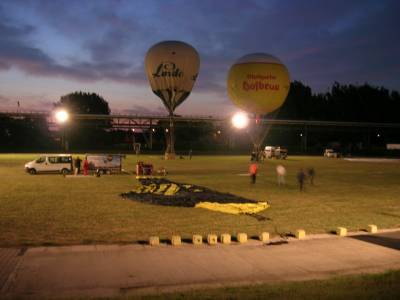 Das Bild zeigt den Ballonstartplatz Bitterfeld. Im Vordergrund liegt ein Heißluftballon füllbereit. In der Bildmitte stehen zwei Gasballone. Während der rechte gelb-weiße Ballon mit der roten Aufschrift Stuttgarter Hofbräu bereits fertig gefüllt ist, ist der blau-weiße Ballon links mit der Aufschrift Linde erst zur Hälfte gefüllt. Hinter den Ballonen ist die Rohrbrücke mit den Gasleitungen zu sehen.